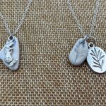 "Two silver chain necklaces displayed on a burlap-like fabric, one featuring a silver mermaid charm with a smooth stone, the other featuring a small engraved stone and an oval silver leaf charm.